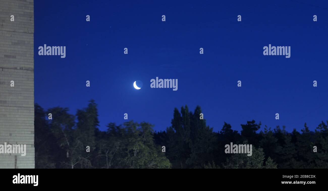 Aschenes Licht des Mondes im hellen Schein der abnehmenden Mondsichel über einem dunklen Wald, gegen einen klaren Nachthimmel, Ecke des Gebäudes links Stockfoto