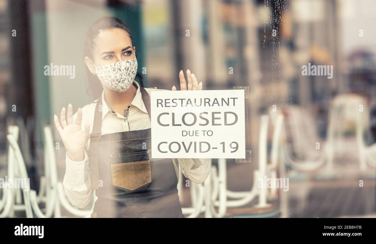 Kellnerin in einem geschlossenen Restaurant wegen Covid-19 trägt Gesichtsmaske, die aus dem Fenster schaut. Stockfoto