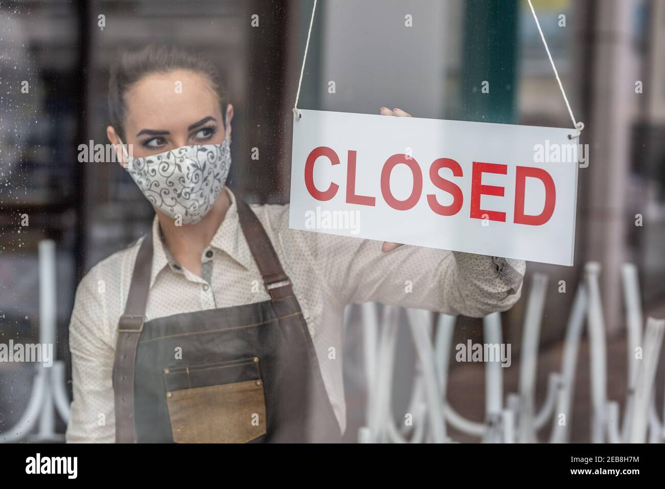 Unglückliche Kellnerin trägt Gesichtsmaske dreht ein Zeichen in einer Bar wegen Coronavirus geschlossen. Stockfoto