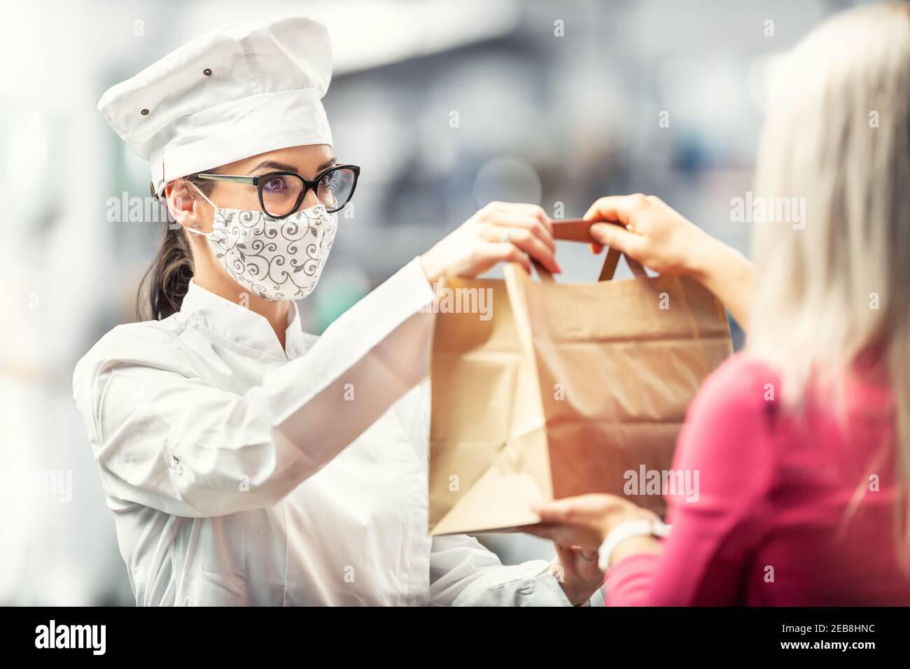 Koch trägt Hut, Gläser und Gesichtsmaske Hände eine Papiertasche mit Lieferung Lebensmittel an einen Kunden. Stockfoto