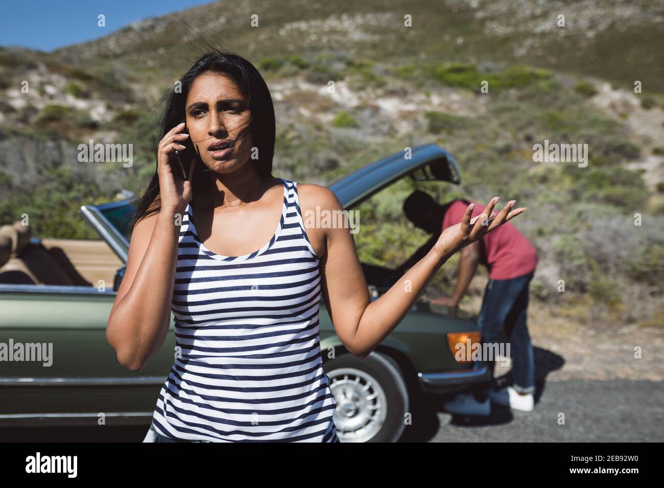 Mixed Race Frau im Gespräch mit Smartphone auf der Straße neben stehen Abgebrochenes Auto mit offener Motorhaube Stockfoto