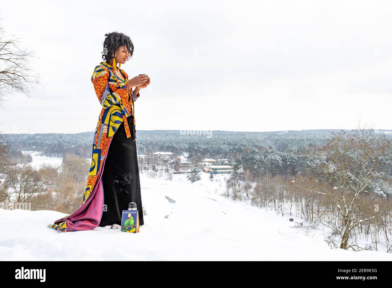 Schönes Mädchen mit bunten Kleid trinken heißen Kaffee oder Tee Auf dem Schnee im Winter mit Waldbäumen bedeckt Schnee und gefrorener Fluss im Hintergrund Stockfoto