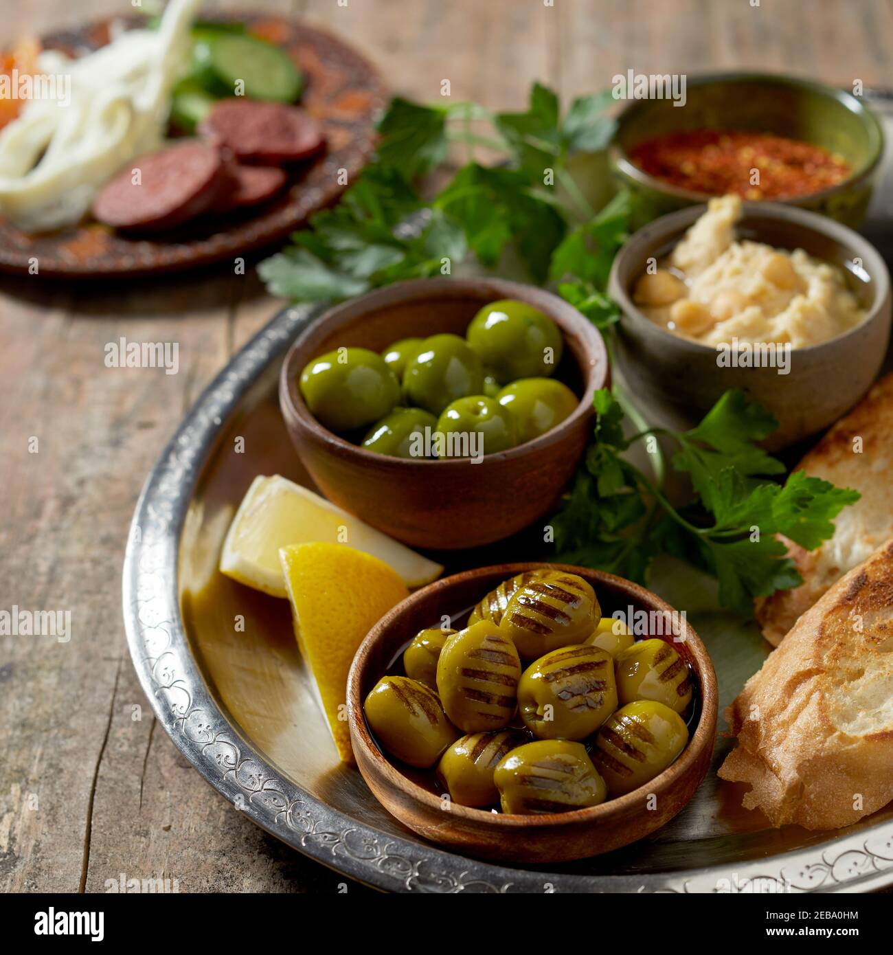 Gesundes mediterranes Frühstück mit gegrillten grünen Oliven, Kichererbsenpaste und geröstetem Baguette auf einem Metalltablett über einem rustikalen Tisch serviert Stockfoto