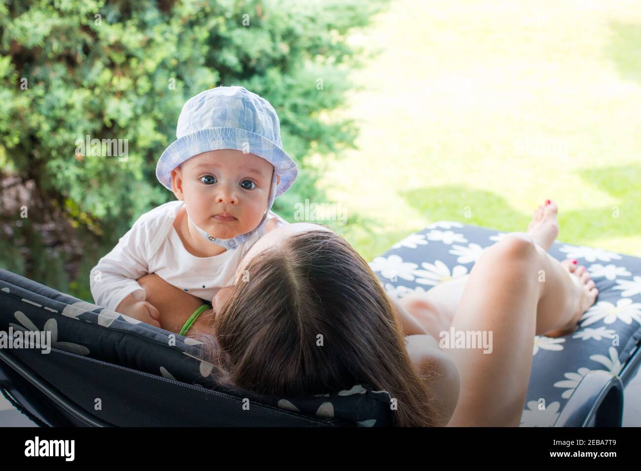 Schöne Mutter hält neugierige Baby Junge in Hut, während legt sich auf Blumenmuster Liegestuhl, genießen Mutterschaft draußen Stockfoto