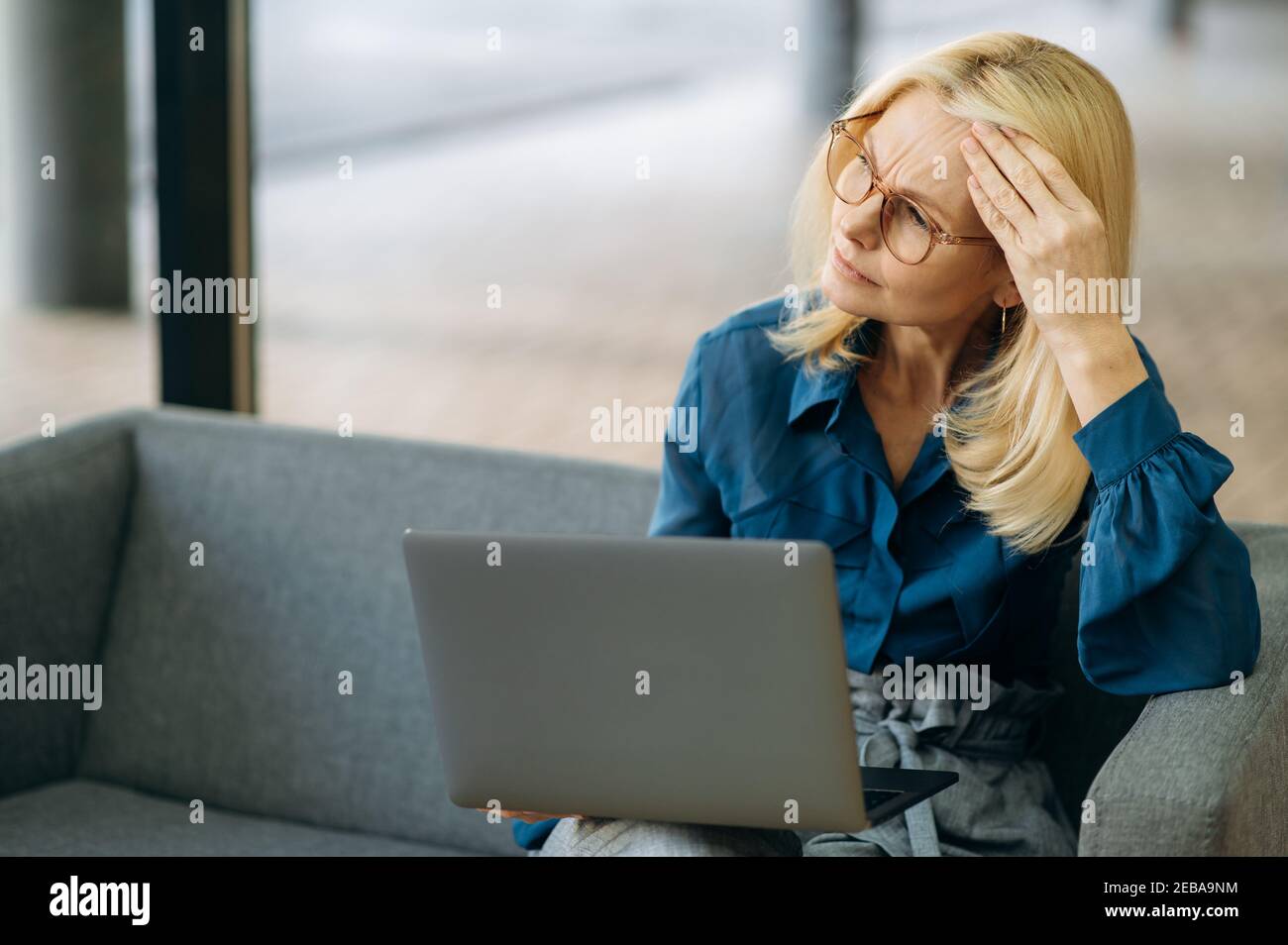 Ältere Erwachsene Geschäftsfrau sitzt auf der Couch, mit Laptop. Erschöpfte weibliche Mitarbeiterin hat eine schreckliche Kopfschmerzen, Gefühl schlecht nach angespanntem Brainstorming, Überarbeit Konzept Stockfoto