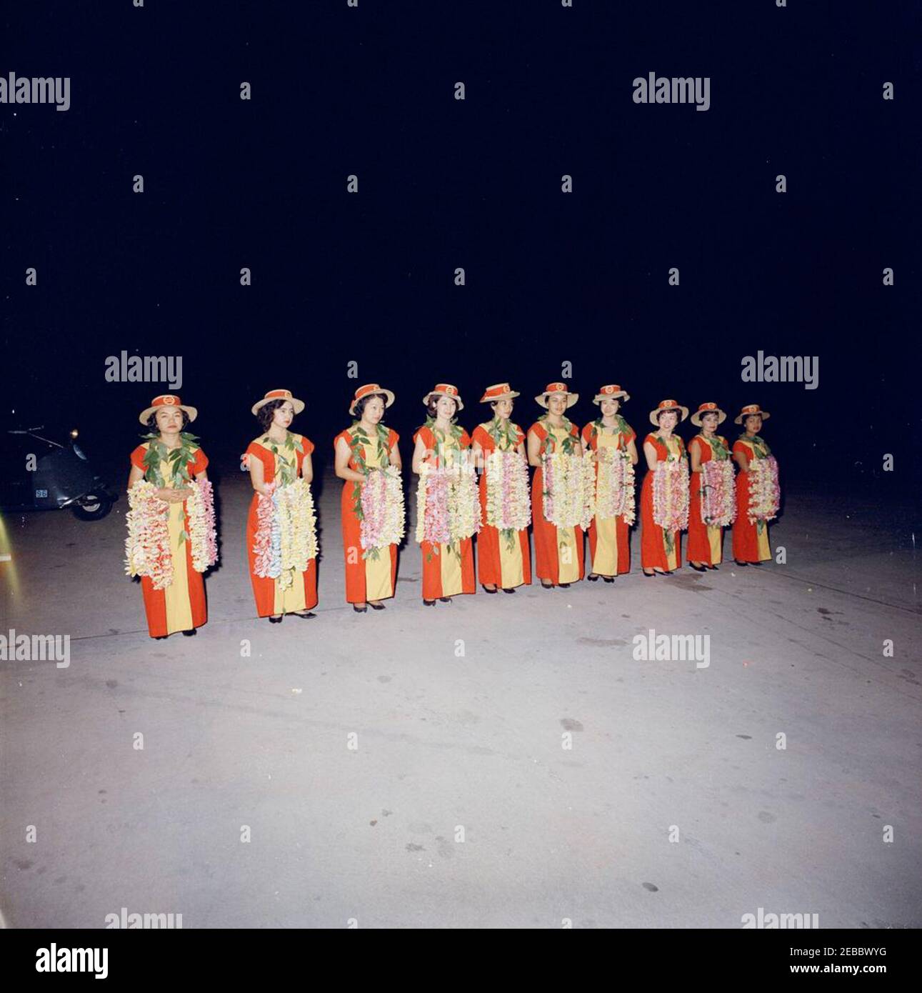 Reise in die westlichen Staaten: Honolulu, Hawaii, Ankunft am Honolulu International Airport, 9:10pm Uhr. Zehn Frauen (in roten und gelben Kleidern mit passenden Hüten und mit Blumenleis) stehen in einer Reihe am Honolulu International Airport in Honolulu, Hawaii. Stockfoto