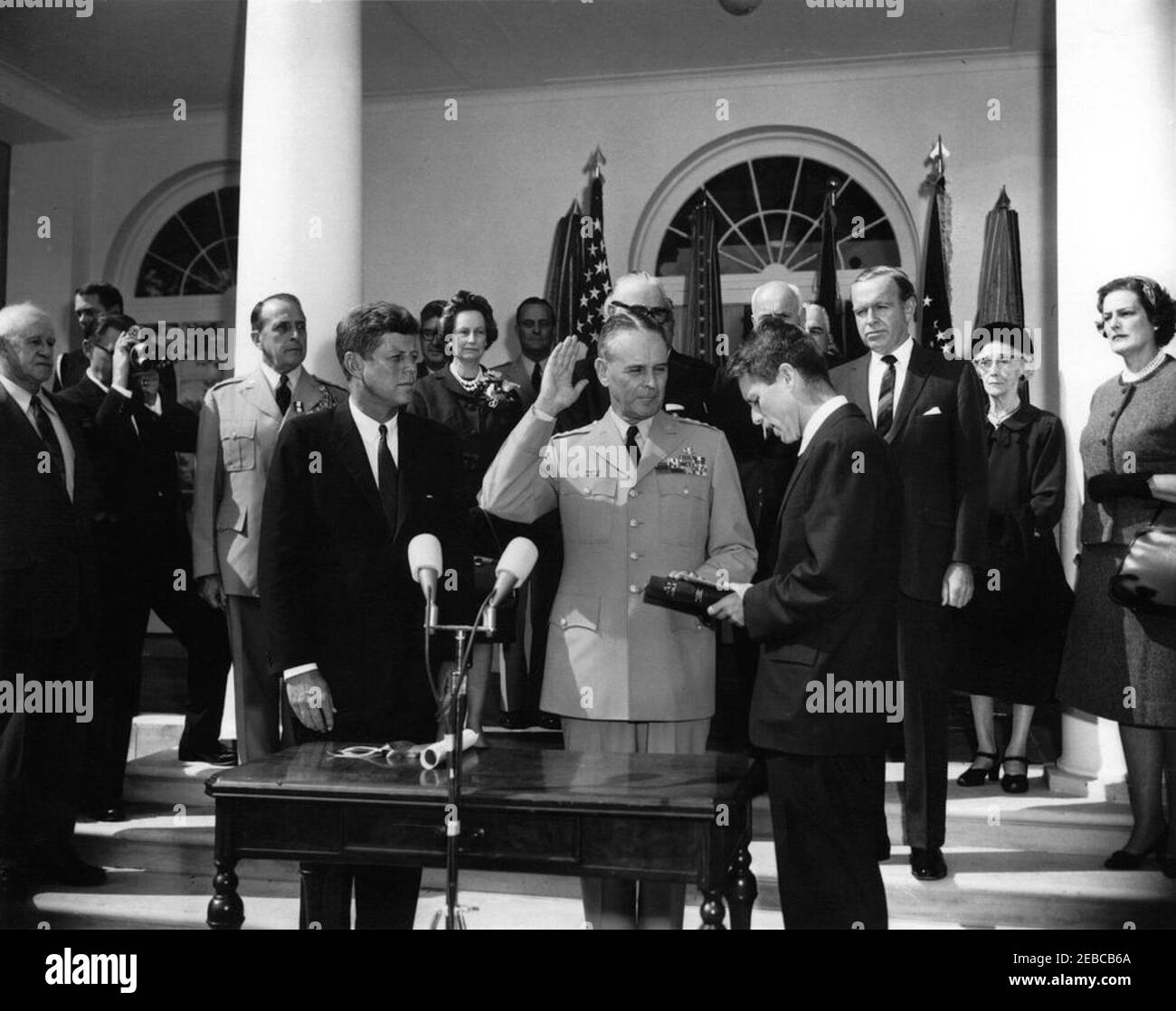 Vereidigung, General Maxwell Taylor, Chairman, JCS, 12:00pm Uhr. Präsident John F. Kennedy (Mitte links) und andere beobachten als General Maxwell D. Taylor (mit rechter Hand angehoben und linke Hand auf der Bibel) wird als Vorsitzender der Joint Chiefs of Staff (JCS) vereidigt; Generalstaatsanwalt Robert F. Kennedy (Mitte rechts, hält Bibel), verwaltet den Amtseid. Blick von den Stufen der West Wing Colonnade, beginnend ganz hinten (von links nach rechts): Naval Aide to the President, Captain Tazewell T. Shepard, Jr.; General Omar Bradley; unidentifizierter Filmfotograf; Outgoing Chai Stockfoto