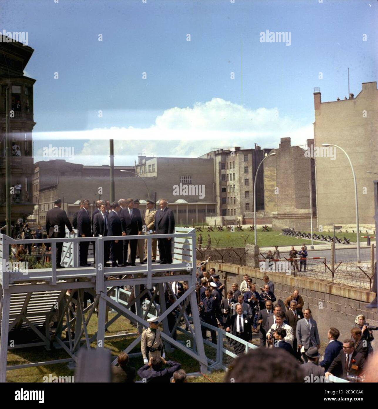 Europareise: Deutschland, West-Berlin: Präsident Kennedy am Checkpoint Charlie, 12:05pm Uhr. Präsident John F. Kennedy blickt von einer erhöhten Plattform am Checkpoint Charlie entlang der Berliner Mauer in West-Berlin (Bundesrepublik) auf Ost-Berlin, DDR (Demokratische Republik). Ebenfalls im Bild: Bundeskanzler Konrad Adenauer; Militärhilfe an Präsident Kennedy, General Chester V. Clifton; Kommandierender General der Berliner Brigade, Generalmajor Frederick O. Hartel; US-Staatssekretär, Dekan Rusk; Minister für All-German Affairs, Dr. Rainer Barzel; US-Protokollchef, Stockfoto