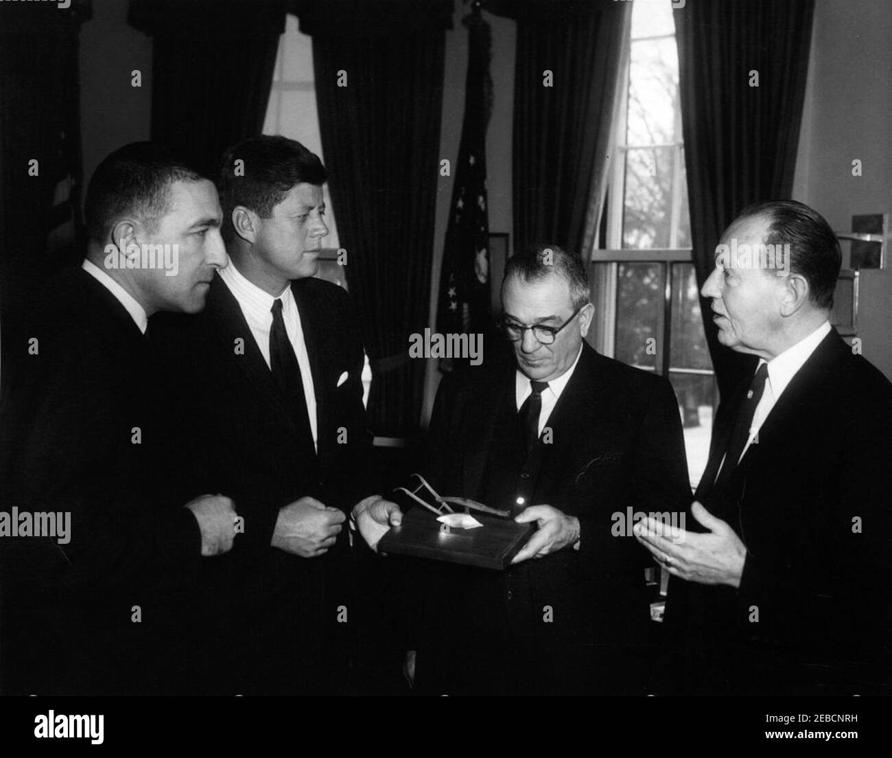 Gedenken an die Hundertjahrfeier der Homestead Act, 12:08pm. Präsident John F. Kennedy nimmt ein Geschenk als Teil einer Gedenkfeier zum hundertjährigen Bestehen des Homestead Act, Oval Office, White House, Washington, D.C. Willis G. Scholl, Präsident des Farm Equipment Institute, und C.R. Gutermuth, Vizepräsident des Wildlife Management Institute, übergibt dem Präsidenten ein fußlanges Modell eines Stahlpfluges. Innenminister Stewart Udall steht auf der linken Seite. Stockfoto