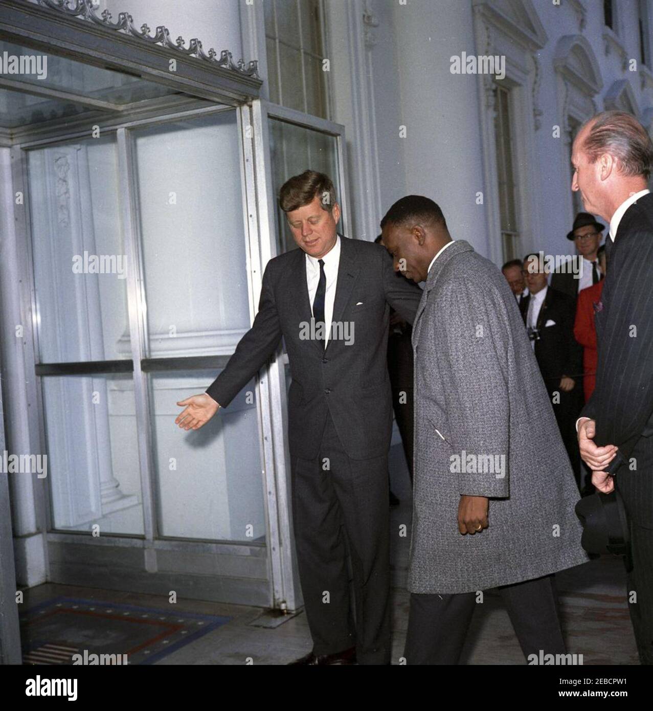 Mittagessen zu Ehren von Cyrille Adoula, Premierminister der Republik Kongo (Leopoldville), 1:00pm Uhr. Präsident John F. Kennedy steht beiseite, als der Premierminister der Republik Kongo (Leopoldville), Cyrille Adoula, das Weiße Haus betritt; Premierminister Adoula besuchte das Weiße Haus, um zu seinen Ehren ein Mittagessen zu besuchen. Chef des Protokolls, Angier Biddle Duke, steht ganz rechts. Mitglieder der Presse stehen im Hintergrund. North Portico, White House, Washington, D.C. Stockfoto
