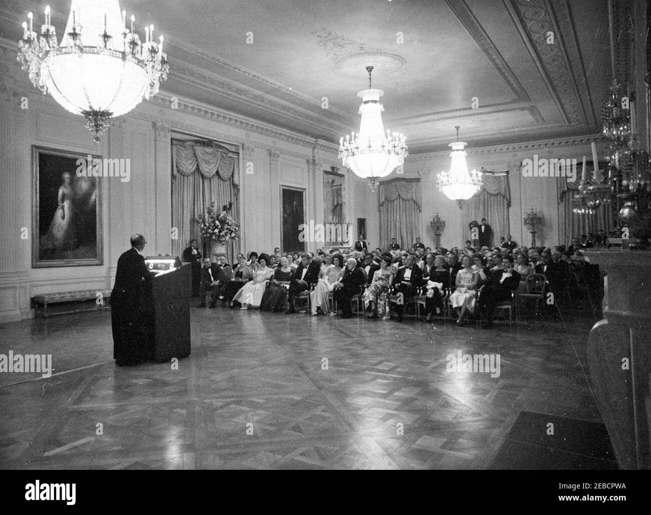 Abendessen zu Ehren der Nobelpreisträger, 8:00pm Uhr. Präsident John F. Kennedy und andere treffen sich im East Room, um bei einer Lesung des Schauspielers Frederic March von Auszügen aus den Werken der späten amerikanischen Nobelpreisträger während eines Abendessens zu Ehren der Nobelpreisträger aus der westlichen Hemisphäre teilzunehmen; ein nicht identifizierter Mann (links) steht am Rednerpult. Platz in der ersten Reihe (L-R): Vizepräsident Lyndon B. Johnson; Katherine Tupper Marshall (Witwe des Nobelpreisträgers und ehemaligen Staatssekretärs, General George C. Marshall); Lady Bird Johnson; Nobelpreisträger, Pearl S. Buck; Präsident Kennedy; erste Stockfoto