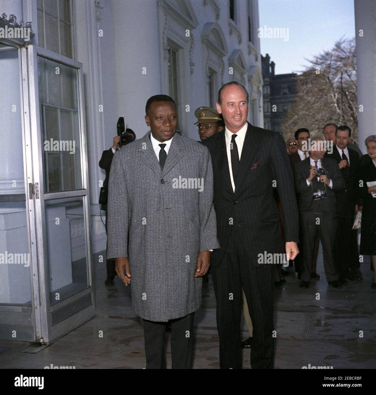 Mittagessen zu Ehren von Cyrille Adoula, Premierminister der Republik Kongo (Leopoldville), 1:00pm Uhr. Der Premierminister der Republik Kongo (Leopoldville), Cyrille Adoula, steht mit dem Protokollführer Angier Biddle Duke (rechts), nach Adoulau2019s Ankunft im Weißen Haus für ein Mittagessen zu seinen Ehren. Ein nicht identifizierter kongolesischer Offizier steht hinten; Washington-Korrespondent für die Guy Gannet Publishing Company of Maine, May Craig (ganz rechts), und andere Mitglieder der Presse stehen rechts. North Portico, White House, Washington, D.C. Stockfoto