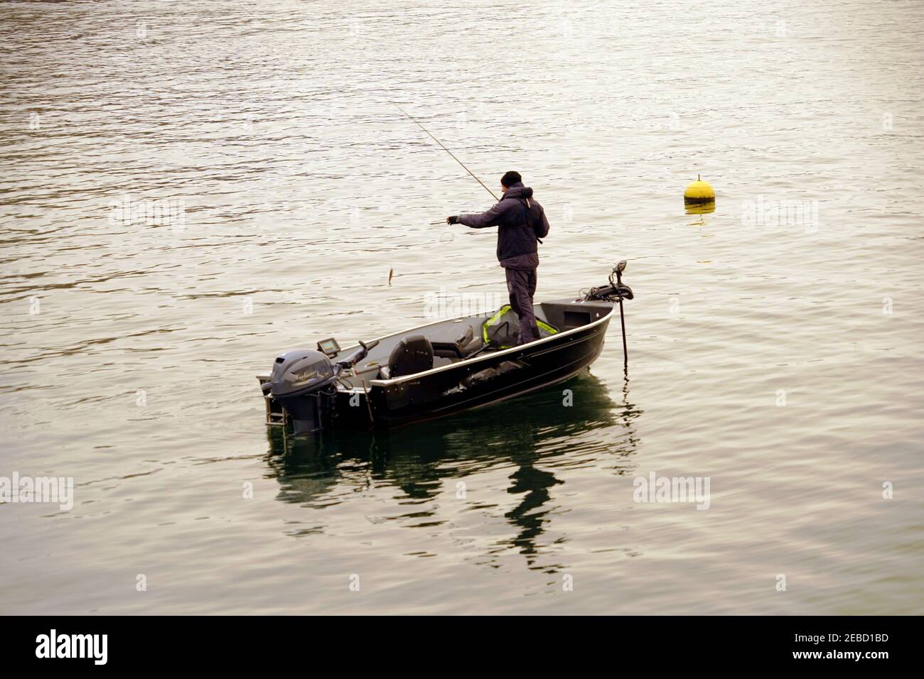 Ein Hobbyfischer, der im Winter mit einem Fisch aus dem Zürichsee den Haken fischt. Er steht in einem Boot und es gibt eine gelbe Boje auf dem Wasser. Stockfoto