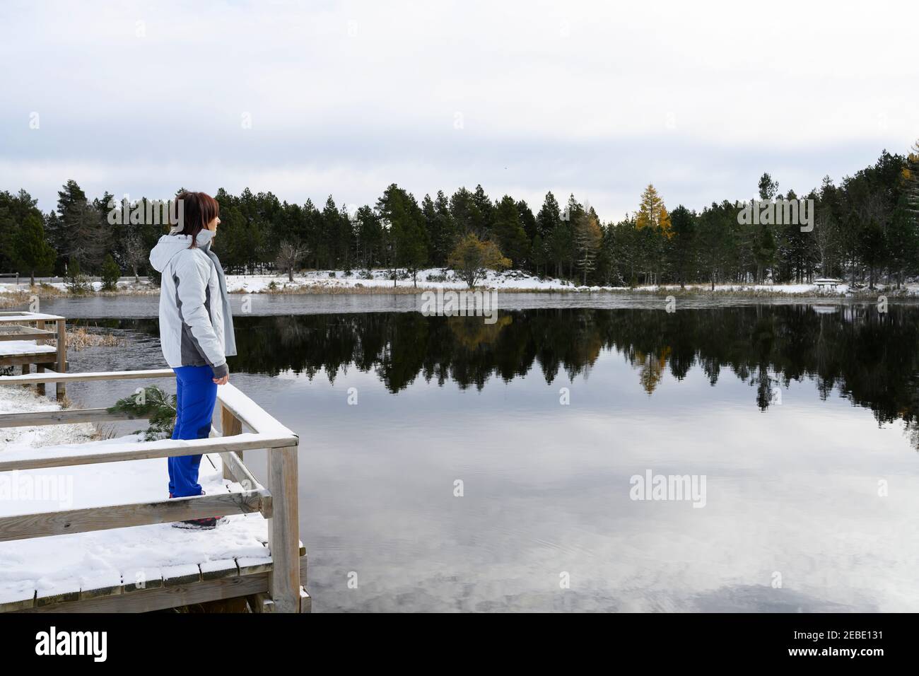 Frau, die auf einer verschneiten Holzbrücke steht und auf eine gefrorene schaut see Stockfoto