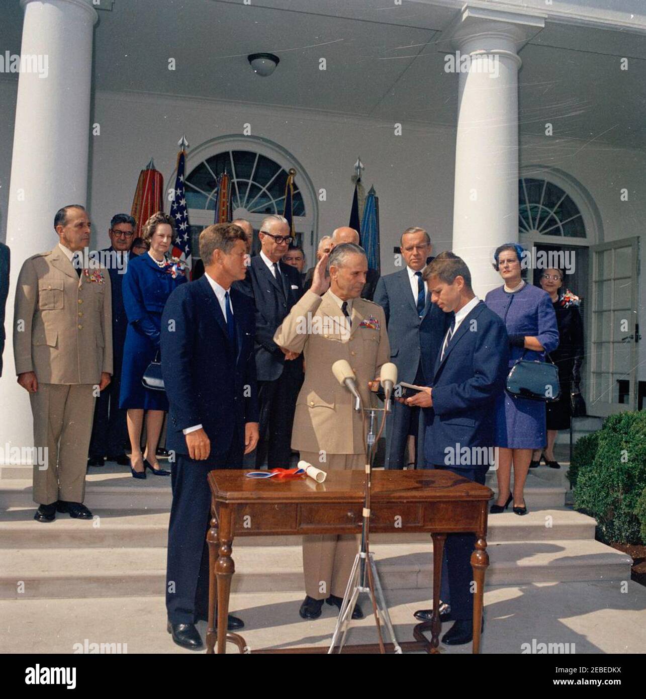 Vereidigung, General Maxwell Taylor, Chairman, JCS, 12:00pm Uhr. Präsident John F. Kennedy (Mitte links) und andere beobachten als General Maxwell D. Taylor (mit rechter Hand angehoben und linke Hand auf der Bibel) wird als Vorsitzender der Joint Chiefs of Staff (JCS) vereidigt; Generalstaatsanwalt Robert F. Kennedy (Mitte rechts, hält Bibel), verwaltet den Amtseid. Blick von den Stufen der West Wing Colonnade (links nach rechts): Scheidender Vorsitzender der JCS, General Lyman L. Lemnitzer; Stabschef der United States Air Force, General Curtis E. LeMay; Lydia Taylor; Admiral Arthur W. Radfor Stockfoto