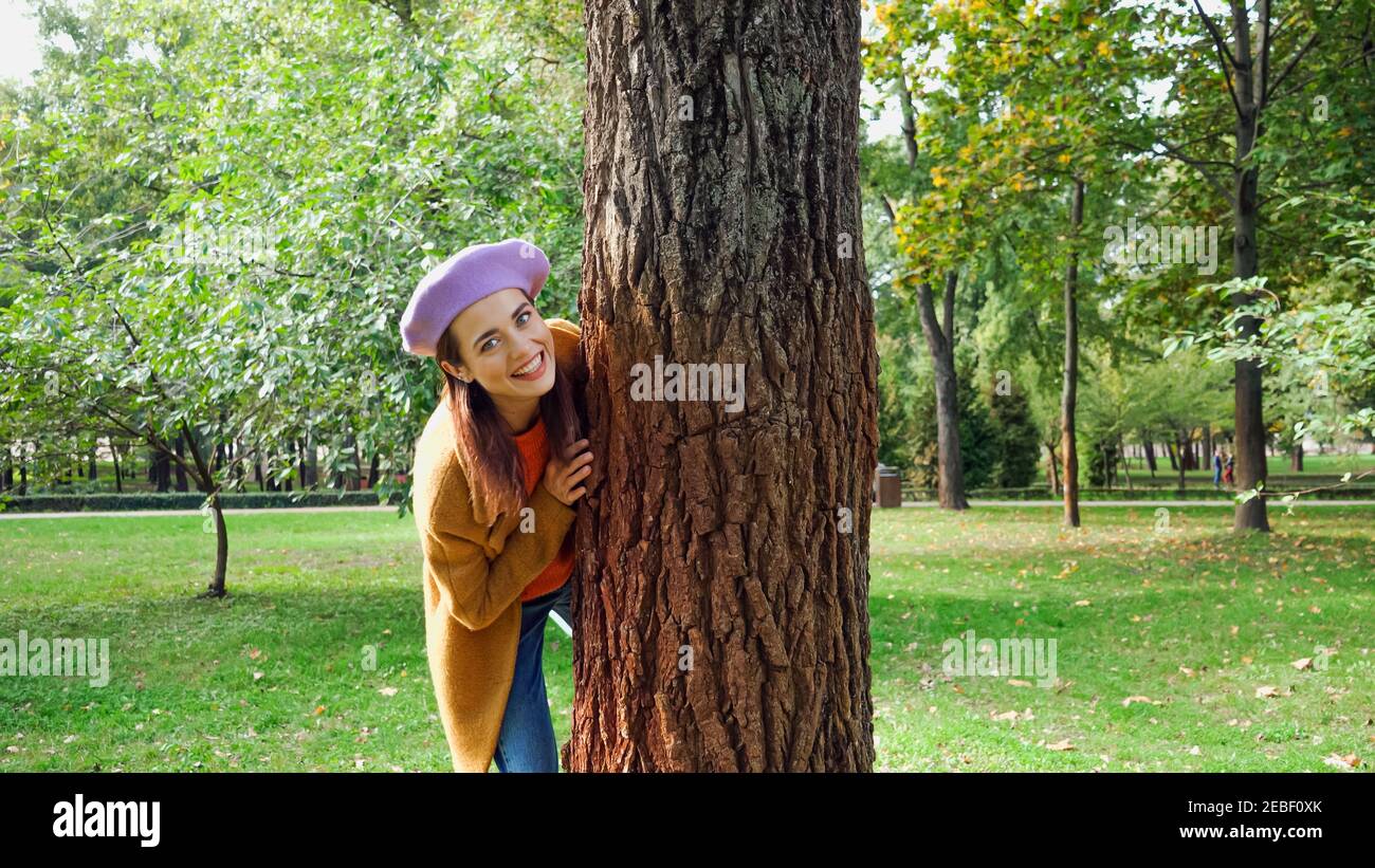 Aufgeregt Frau versteckt hinter Baumstamm im Park beim Blick Bei der Kamera Stockfoto
