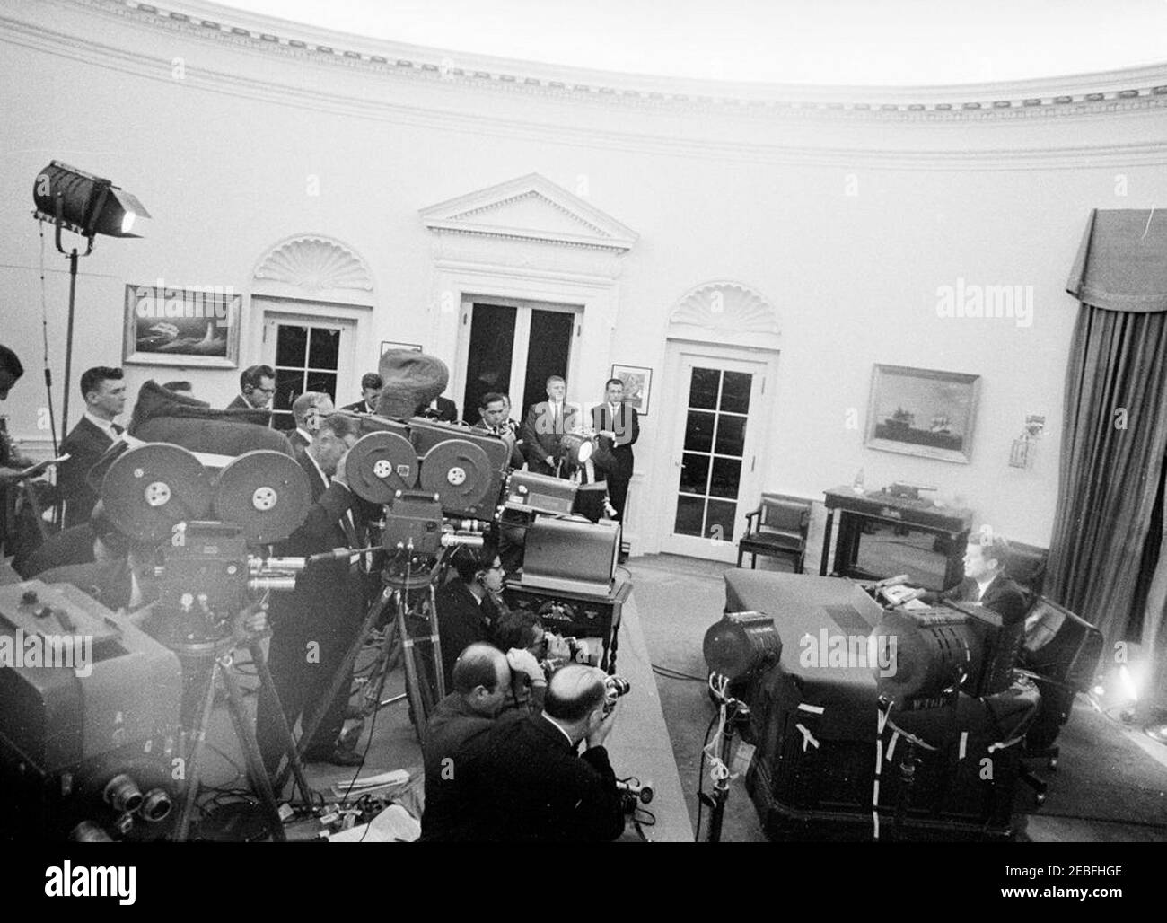 Radio und Fernsehen Bericht an das amerikanische Volk über den Aufbau sowjetischer Waffen in Kuba (Kubakrise), 7:00pm Uhr. Kameraleute Film Präsident John F. Kennedy (ganz rechts, sitzt am Schreibtisch), als er eine Radio-und Fernsehansprache an die Nation in Bezug auf die sowjetische Militärpräsenz Unionu2019s in Kuba. Die Agenten des Geheimdienstes des Weißen Hauses, Dennis Halterman und Gerald u201cJerryu201d Blaine (Arme gefaltet), stehen im Hintergrund, links vom Eingang. Oval Office, White House, Washington, D.C. Stockfoto