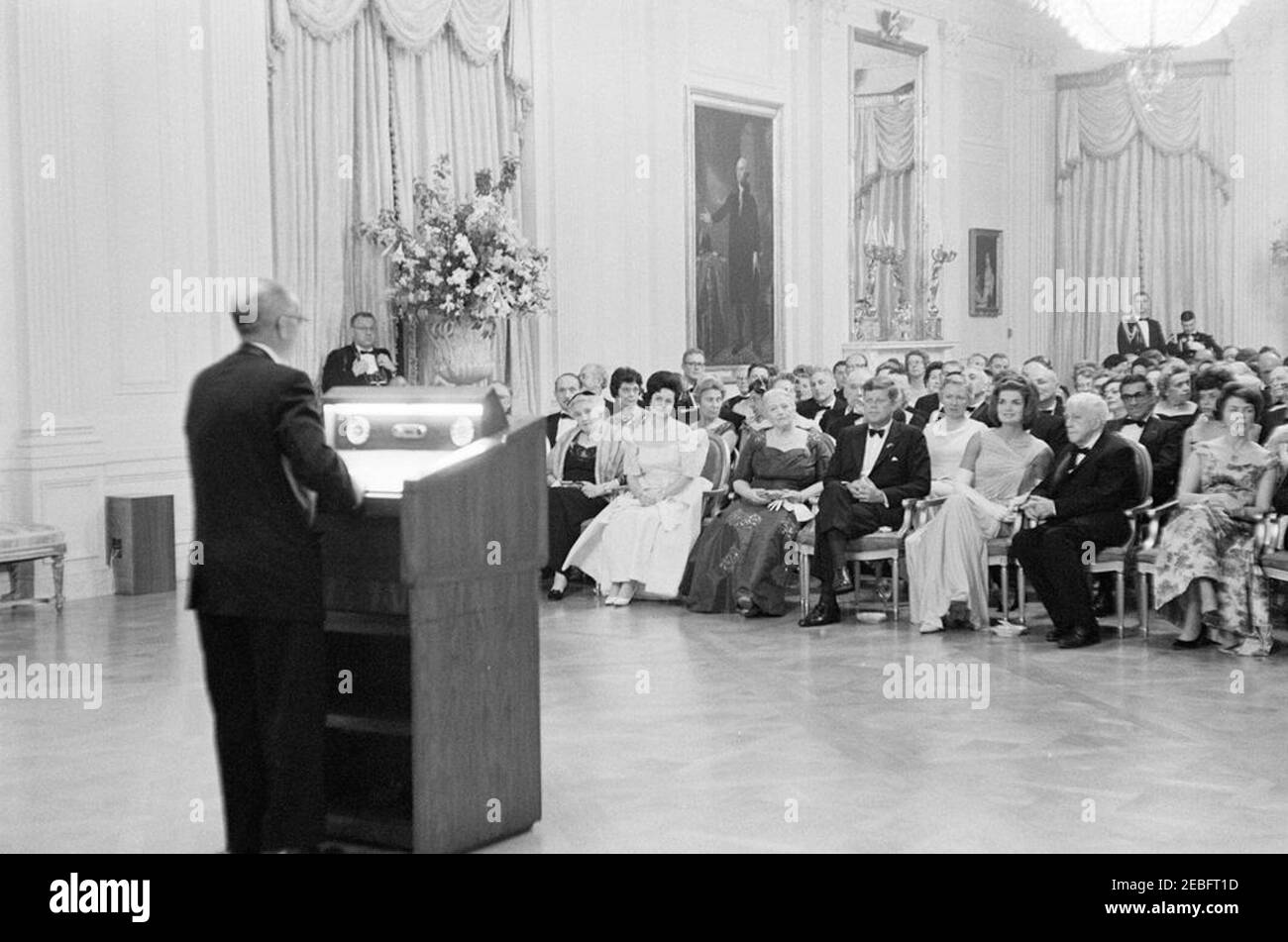 Abendessen zu Ehren der Nobelpreisträger, 8:00pm Uhr. Präsident John F. Kennedy und andere treffen sich im East Room, um bei einer Lesung des Schauspielers Frederic March von Auszügen aus den Werken der späten amerikanischen Nobelpreisträger während eines Abendessens zu Ehren der Nobelpreisträger aus der westlichen Hemisphäre teilzunehmen; ein nicht identifizierter Mann (links) steht am Rednerpult. Platz in der ersten Reihe (L-R): Katherine Tupper Marshall (Witwe des Nobelpreisträgers und ehemaligen Staatssekretärs, General George C. Marshall); Lady Bird Johnson; Nobelpreisträger-Autorin, Pearl S. Buck; Präsident Kennedy; First Lady Jacqueline Kennedy; Dichterin und Stockfoto