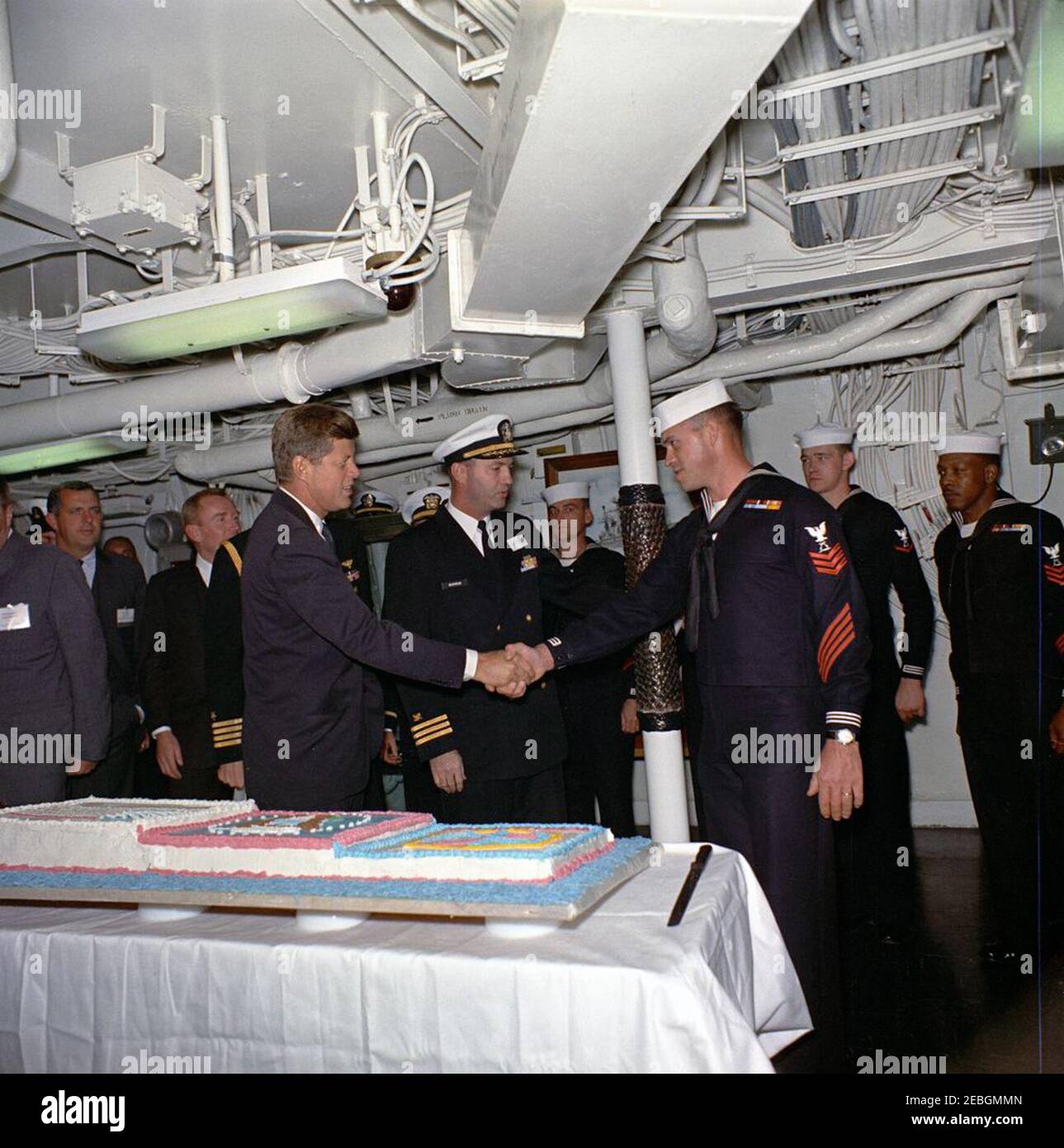 Besuch der Atlantischen Flotte: Präsentation einer Torte an Präsident Kennedy an Bord der USS Northampton (CC-1). Präsident John F. Kennedy (links) schüttelt sich die Hände mit schüttelt die Hände mit Commissary Steward Petty Officer First Class, Horace L. Medders (USN), während einer Kuchenschneidezeremonie zum Gedenken an seinen Besuch an Bord des Kreuzfahrtschiffes USS Northampton (CC-1), Kommandoschiff der US-Atlantikflotte, Auf See vor der Küste von Virginia und North Carolina; Officer Medders war einer der Seeleute, die den Kuchen gebacken. Assistant Secretary of the Treasury James A. Reed (hinten, zweiter von links) und Unterstaatssekretär von Stockfoto