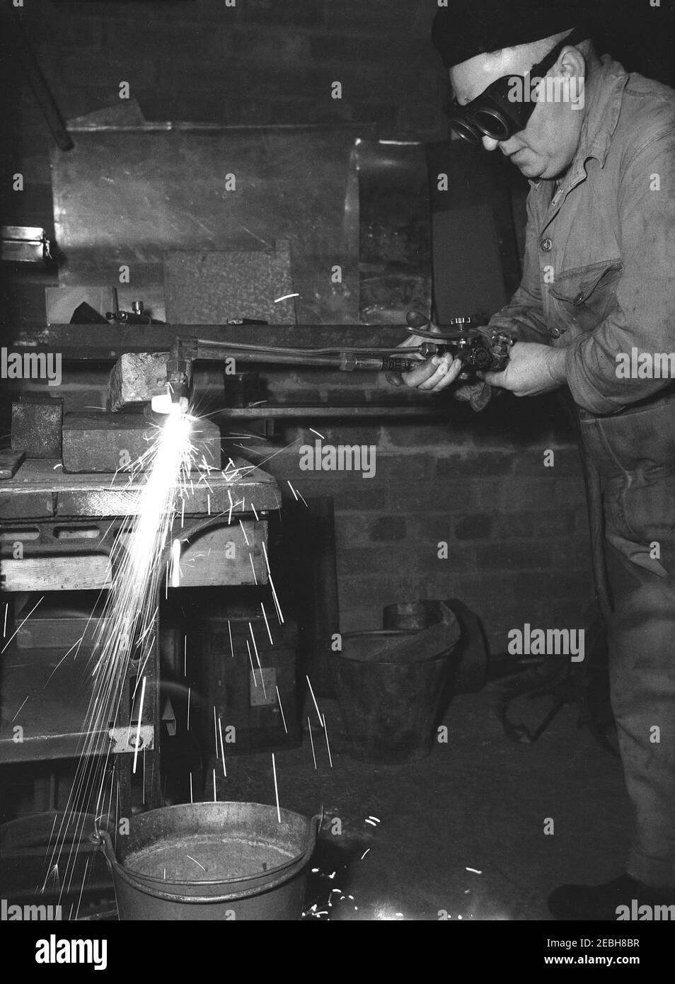 1950s, historisch, ein Handwerker in Overalls und Tragen von Brillen und einer Beere auf seinem Hut in einer kleinen Werkstatt tun Metallarbeiten oder Schweißen. Schweißen ist der Prozess der Verbindung von Metallen. Fusion ist der generische Begriff für die Herstellung von Blechen für die Verbindung von Metallen ähnlicher Zusammensetzungen und Schmelzpunkte. Aufgrund der hohen Schmelzpunkte der Werkstücke entsteht ein Schmelzbad, das sogenannte Schweißbad. Stockfoto