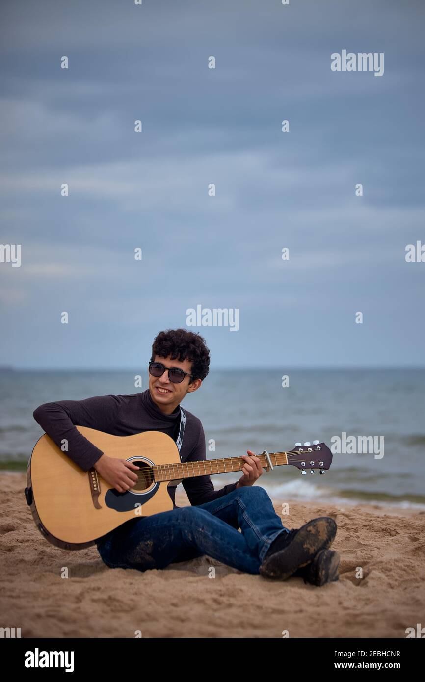 Junge kaukasische Teenager spielen akustische Gitarre am Strand. Sonnenbrillen und dunkle Kleidung. Stockfoto