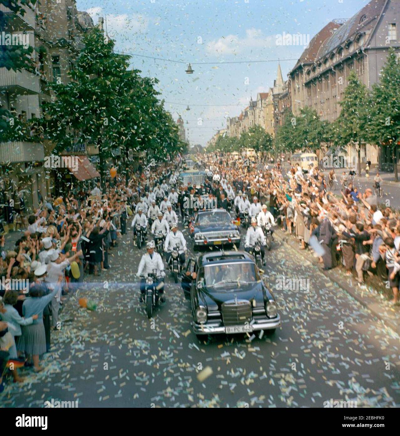 Europa-Reise: Deutschland, West-Berlin: Präsident Kennedy im Autokat mit Willie Brandt, Bürgermeisterin von West-Berlin, und Konrad Adenauer, Bundeskanzler von West-Deutschland. Präsident John F. Kennedy (stehend im zweiten Auto) winkt von der Präsidentenlimousine (Lincoln-Mercury Continental Cabrio) zu Menschenmengen, während seine Autokolonne durch West-Berlin, Westdeutschland (Bundesrepublik) reist. Mit dem Präsidenten: Bürgermeister von West-Berlin, Willy Brandt; Bundeskanzler Konrad Adenauer. Die Leute säumen die Straße und werfen Konfetti. [Der blaue Streifen auf dem Bild entspricht dem Negativ.] Stockfoto