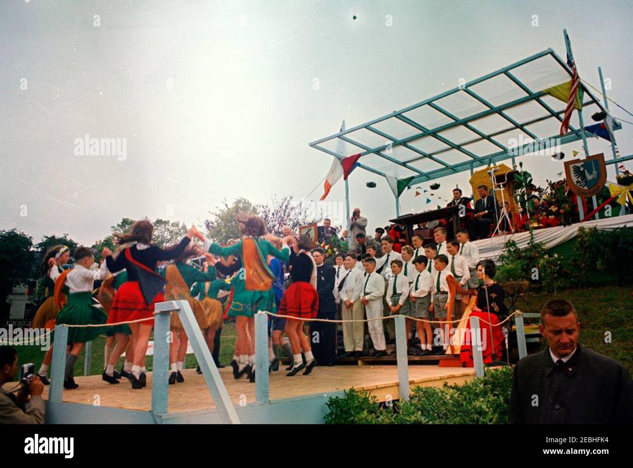 Europareise: Irland, Galway: Präsident Kennedy am Eyre Square, Bemerkungen und Abreise, 11:55am Uhr. Präsident John F. Kennedy (oben rechts), Bürgermeister von Galway, Alderman Patrick D. Ryan (links von Präsident Kennedy), Und andere hochrangige Gäste beobachten von der oberen Plattform aus, wie Kinder von lokalen Schulen (unten links) während einer Begrüßungszeremonie für den Präsidenten auf dem Eyre Square in Galway, Irland, einen traditionellen irischen Tanz vorführen; Kinder des Patrizier Brothers Choir stehen rechts von den Tänzern, mit Harfenistin Ruth Bradley (sitzend, mit rotem Rock). Ebenfalls abgebildet (in der oberen Plattform sitzend, Stockfoto