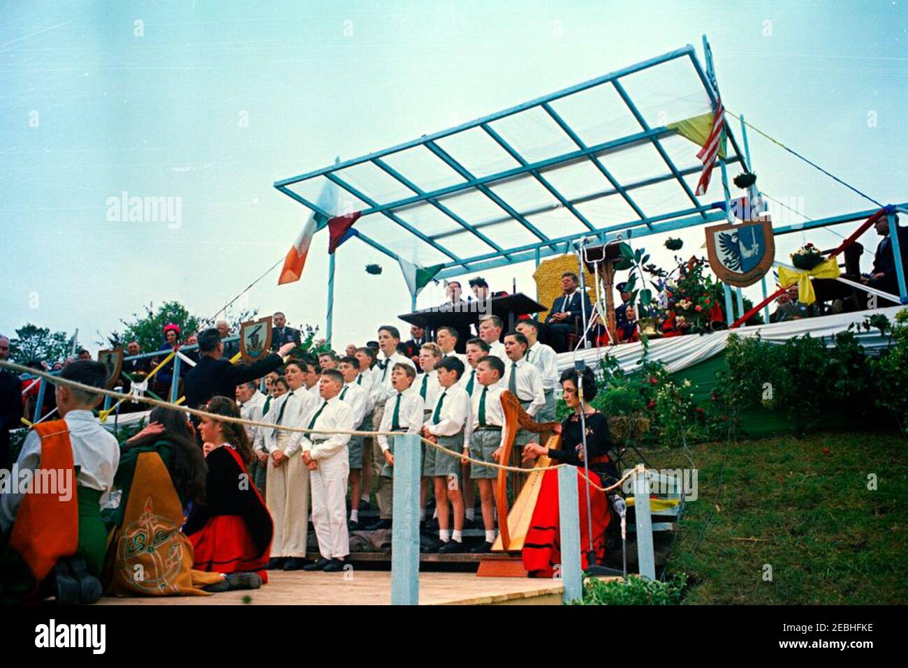 Europareise: Irland, Galway: Präsident Kennedy am Eyre Square, Bemerkungen und Abreise, 11:55am Uhr. Präsident John F. Kennedy (oben rechts), Bürgermeister von Galway, Alderman Patrick D. Ryan (links von Präsident Kennedy), Und andere hochrangige Gäste beobachten von der oberen Plattform aus, wie Kinder des Patrizier Brothers Choir (unten in der Mitte) singen, begleitet von der Harfenistin Ruth Bradley, während einer Begrüßungszeremonie für den Präsidenten auf dem Eyre Square in Galway, Irland; unten links sitzt eine Gruppe von Kindern aus lokalen Tanzschulen. Ebenfalls abgebildet (auf der oberen Plattform, unter der irischen Flagge): United St Stockfoto