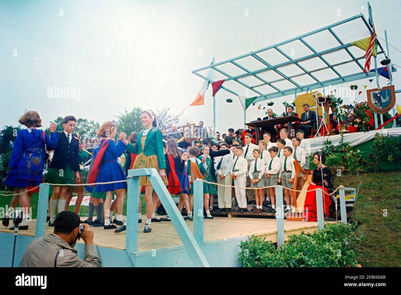 Europareise: Irland, Galway: Präsident Kennedy am Eyre Square, Bemerkungen und Abreise, 11:55am Uhr. Präsident John F. Kennedy (oben rechts), Bürgermeister von Galway, Alderman Patrick D. Ryan (links von Präsident Kennedy), Und andere hochrangige Gäste beobachten von der oberen Plattform aus, wie Kinder von lokalen Schulen (unten links) während einer Begrüßungszeremonie für den Präsidenten auf dem Eyre Square in Galway, Irland, einen traditionellen irischen Tanz vorführen; Kinder des Patrizier Brothers Choir (rechts von den Tänzern) singen, begleitet von der Harfenistin Ruth Bradley (sitzend, mit rotem Rock). Auch abgebildet (sitzt in der Gruppe Stockfoto