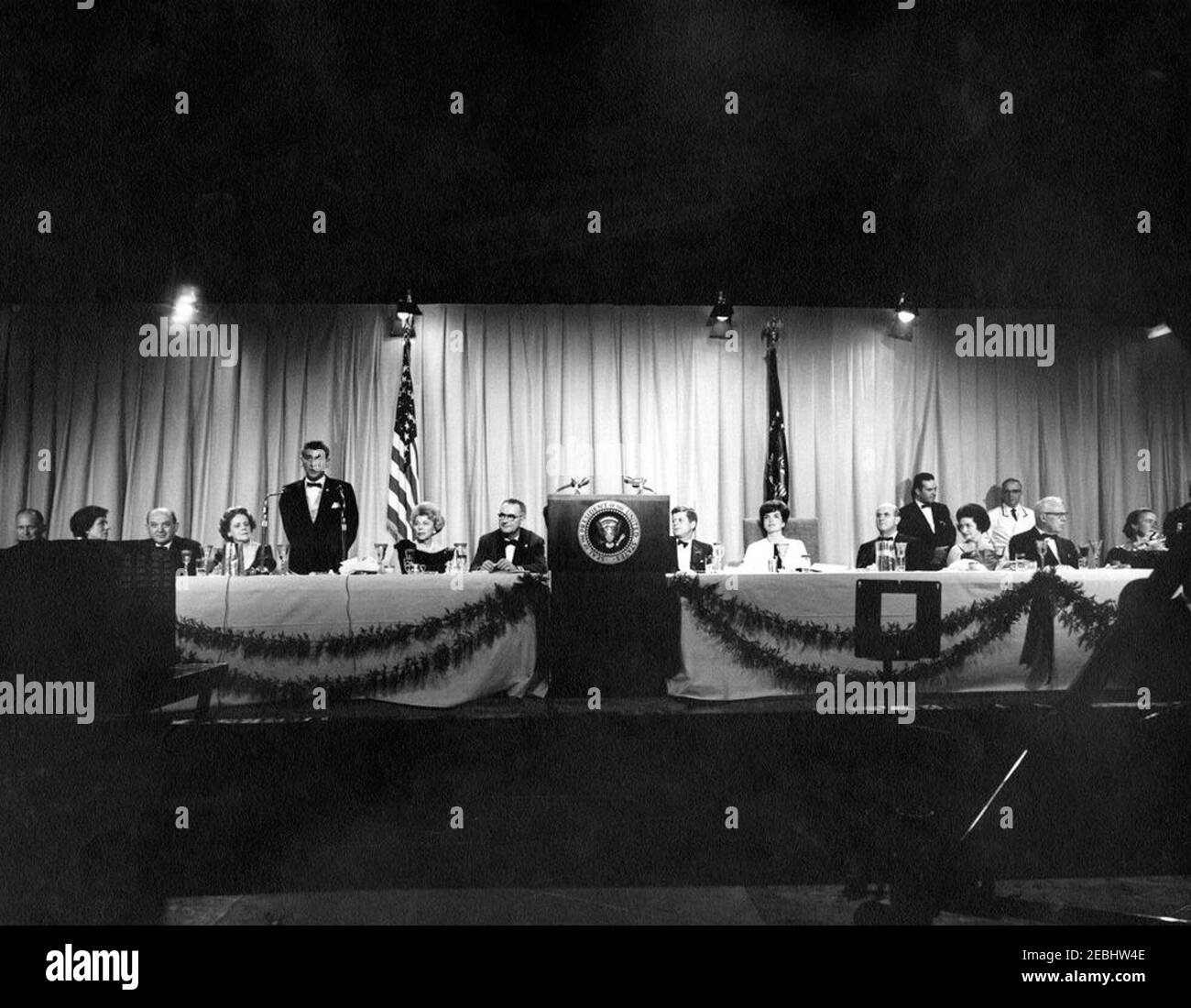 National Cultural Center Fonds-Raising Benefit, 9:23pm. Der Zeremonienmeister, Komponist und Dirigent Leonard Bernstein (links), spricht bei einem Fundraising-Dinner und Closed-Circuit-Telecast von u0022An American Pageant of the Artsu0022 für das National Cultural Center; Präsident John F. Kennedy und First Lady Jacqueline Kennedy nahmen am Dinner Teil. Am Haupttisch (L-R): Finanzminister, C. Douglas Dillon; nicht identifiziert; Staatssekretär, Dean Rusk; Nina Warren, Ehefrau von Earl Warren, Oberrichter des Obersten Gerichtshofs; Herr Bernstein; Vorsitzender des Dinnerkomitees des Nationalen C Stockfoto