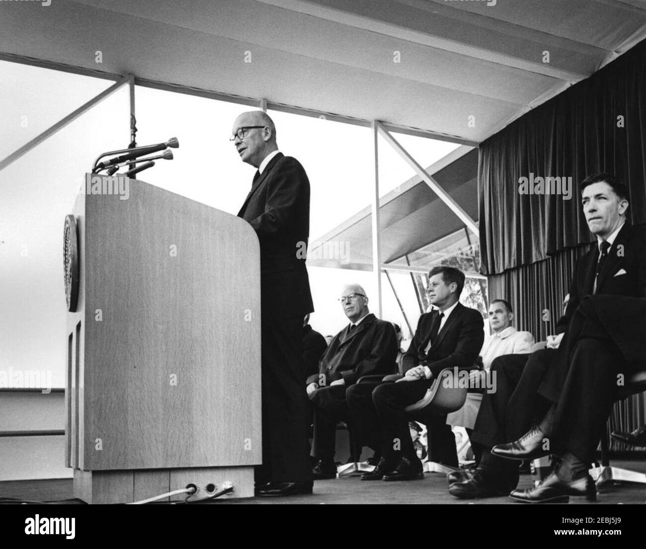 Dedication Ceremonies, Dulles International Airport, 11:12am Uhr. Der ehemalige Präsident, General Dwight D. Eisenhower (am Rednerpult), hält bei der Einweihungszeremonie für den internationalen Flughafen Dulles, benannt nach dem verstorbenen Außenminister John Foster Dulles, eine Rede. Sitzt auf speakersu2019 Plattform (L-R): Chef Richter des Obersten Gerichtshofs, Earl Warren; Präsident John F. Kennedy; White House Secret Service Agent, Floyd Boring (im Rücken); Administrator der Federal Aviation Agency (FAA), Najeeb Halaby. Chantilly, Virginia. Stockfoto