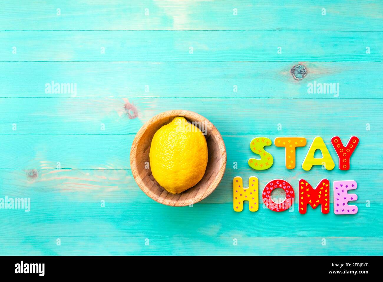 Wörter BLEIBEN ZU HAUSE und gelbe Zitrone in Holzschüsselteller auf türkisfarbenem Holz Hintergrund. Text BLEIBEN SIE HEIMAT von Holzbuchstaben und Obst in Platte Draufsicht. Kopieren Stockfoto