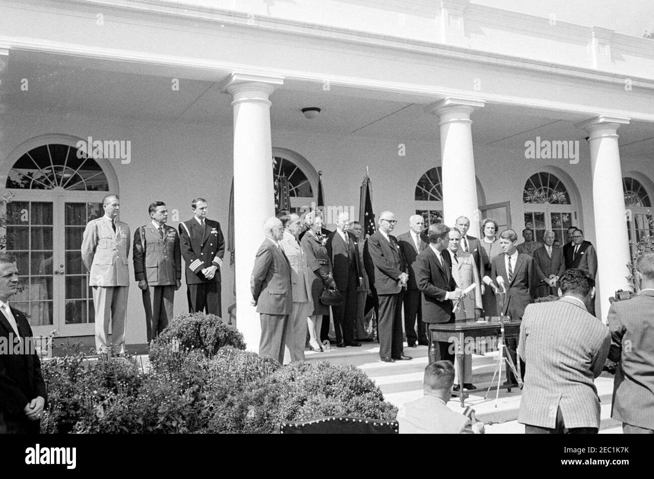 Vereidigung, General Maxwell Taylor, Chairman, JCS, 12:00pm Uhr. Präsident John F. Kennedy (Mitte rechts, an Mikrofonen) hält Bemerkungen während der Vereidigung von General Maxwell D. Taylor als Vorsitzender der Joint Chiefs of Staff (JCS); General Taylor und Generalstaatsanwalt Robert F. Kennedy stehen rechts von Präsident Kennedy. Blick von den Stufen der West Wing Colonnade, beginnend ganz links (links nach rechts): Military Aide an den Präsidenten, General Chester V. Clifton; Air Force Aide an den Präsidenten, Brigadier General Godfrey T. McHugh; Naval Aide an den Präsidenten, Kapitän Stockfoto