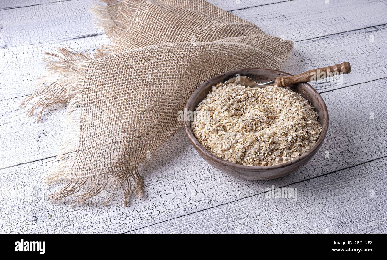 Trockene rohe, ungekochte weiße Quinoa-Flocken in handgefertigter Tonschüssel und Löffel mit Holzgriff auf weiß strukturiertem Holztisch. Leinentischdecke auf si Stockfoto