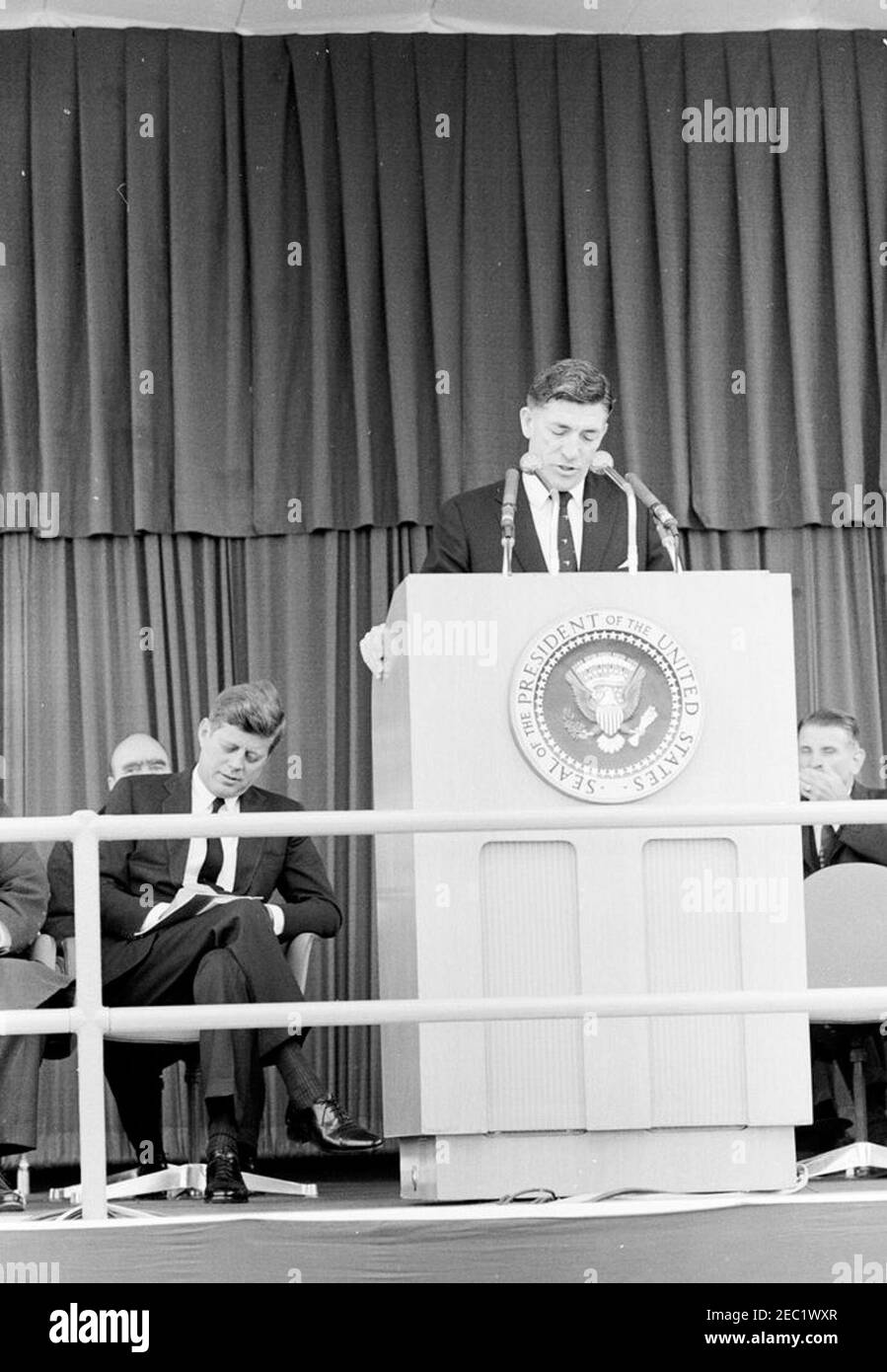 Dedication Ceremonies, Dulles International Airport, 11:12am Uhr. Der Administrator der Federal Aviation Agency (FAA), Najeeb Halaby (am Rednerpult), hält bei der Einweihungszeremonie für den internationalen Flughafen Dulles, benannt nach dem verstorbenen Staatssekretär John Foster Dulles, Rede; Präsident John F. Kennedy (schriftlich) sitzt links. Der stellvertretende Administrator der FAA, Generalleutnant Harold W. Grant (meist versteckt), sitzt hinter Präsident Kennedy; der Vertreter Oren Harris (Arkansas) sitzt ganz rechts. Chantilly, Virginia. Stockfoto