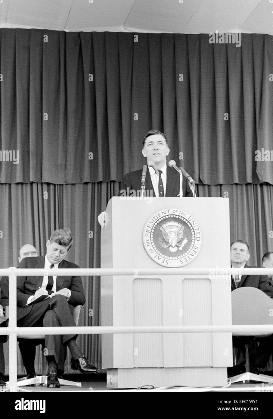 Dedication Ceremonies, Dulles International Airport, 11:12am Uhr. Der Administrator der Federal Aviation Agency (FAA), Najeeb Halaby (am Rednerpult), hält bei der Einweihungszeremonie für den internationalen Flughafen Dulles, benannt nach dem verstorbenen Staatssekretär John Foster Dulles, Rede; Präsident John F. Kennedy (schriftlich) sitzt links. Der stellvertretende Administrator der FAA, Generalleutnant Harold W. Grant (meist versteckt), sitzt hinter Präsident Kennedy; der Vertreter Oren Harris (Arkansas) sitzt ganz rechts. Chantilly, Virginia. Stockfoto