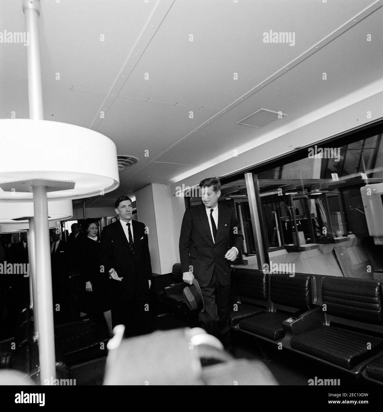 Dedication Ceremonies, Dulles International Airport, 11:12am Uhr. Präsident John F. Kennedy (Mitte, mit Hut) geht durch eine der mobilen Lounges am Dulles International Airport während der Einweihungszeremonie für den Flughafen, der nach dem verstorbenen Staatssekretär John Foster Dulles benannt wurde. Der Verwalter des Luftfahrtbundesamtes (FAA), Najeeb Halaby, und Aline B. Saarinen (Witwe des Architekten für den Flughafen, Eero Saarinen) folgen dahinter. Chantilly, Virginia. Stockfoto