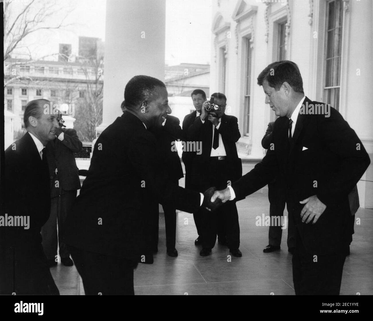 Mittagessen zu Ehren von Sylvanus Olympio, Präsident von Togo, 1:00pm Uhr. Präsident John F. Kennedy (rechts) schüttelt die Hände mit Präsident von Togo, Sylvanus Olympio, bei der Ankunft von Präsident Olympiou2019s im Weißen Haus für ein Mittagessen zu seinen Ehren. Chef des Protokolls, Angier Biddle Duke, steht ganz links; Fotografen, darunter Wochenschau-Fotograf für United Press Movietone, Thomas J. Craven, Sr., stehen im Hintergrund. North Portico, White House, Washington, D.C. Stockfoto