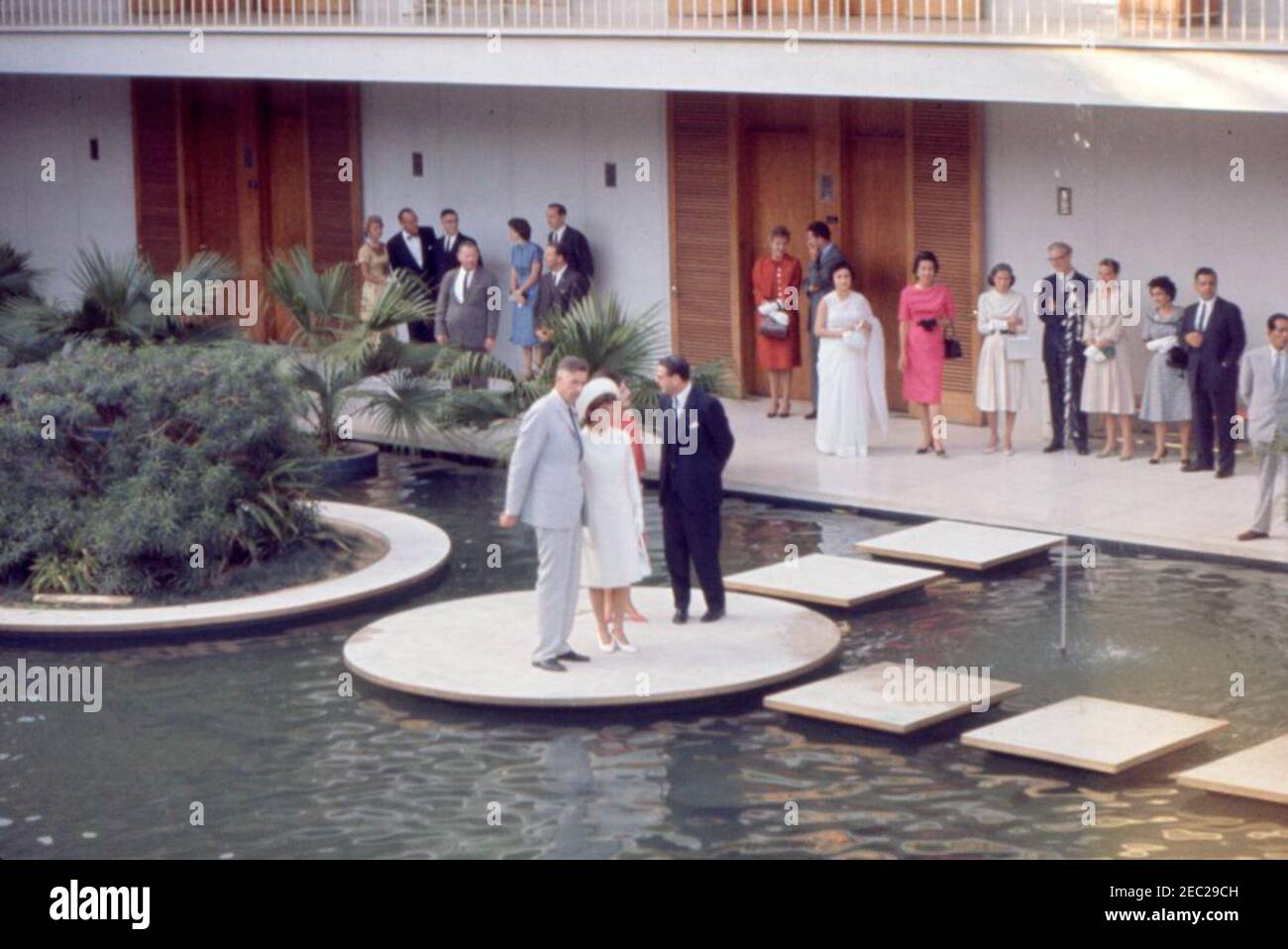 First Lady Jacqueline Kennedyu2019s (JBK) Reise nach Indien und Pakistan: New Delhi, Delhi, Indien, Besuch in US Chancery. First Lady Jacqueline Kennedy (Mitte) blickt auf den Gartenpool in der Kanzlei der Botschaft der Vereinigten Staaten in Neu-Delhi, Indien. Der Botschafter der Vereinigten Staaten in Indien, John Kenneth Galbraith, steht neben Frau Kennedy; die erste Schwester von Ladyu2019s, Prinzessin Lee Radziwill von Polen, steht hinter Frau Kennedy. Kitty Galbraith, die Frau des Botschafters, steht am Gebäude (sechstes von rechts). Stockfoto