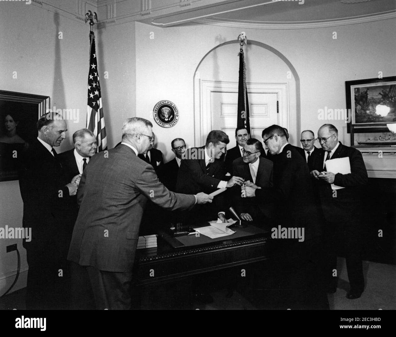 Bill Signing - H.R. 4997 Feed Grain Act, 12:35pm. Präsident John F. Kennedy (am Schreibtisch stehend) reicht dem Landwirtschaftsminister Orville Freeman einen Stift, nachdem er den Feed Grain Act von 1963 unterzeichnet hat. Von links nach rechts: Stellvertretender Landwirtschaftsminister, John P. Duncan, Jr. (vorne, Übergabe einer Kiste an Sekretär Freeman); Vertreter Graham Purcell (Texas); Vertreter W. R. Poage (Texas); Senator Allen J. Ellender (Louisiana); Präsident Kennedy; Präsident der National Farmers Organization, Oren Lee Staley (hinten); Glen J. Talbott von der National Farmers Union; Sekretär Freeman; Executive V Stockfoto