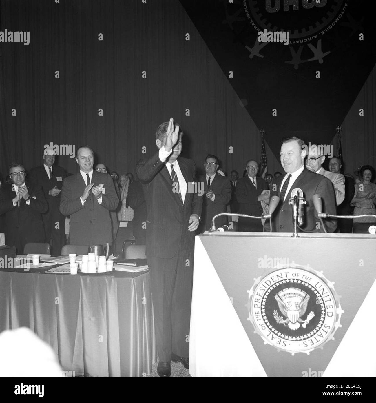 Ansprache vor der United Auto Workers of America (UAW) Convention, Atlantic City, 10:35am Uhr. Präsident John F. Kennedy winkt den Teilnehmern der jährlichen Versammlung der Vereinigten Automobilarbeiter von Amerika (UAW) in der Kongresshalle in Atlantic City, New Jersey. Präsident der UAW, Walter Reuther, steht hinter Mikrofonen; Gouverneur von New Jersey, Richard J. Hughes, steht im Hintergrund (rechts von Präsident Kennedy); Sekretär-Schatzmeister der UAW, Emil Mazey, steht hinter Herrn Reuther. Alle anderen sind nicht identifiziert. Stockfoto