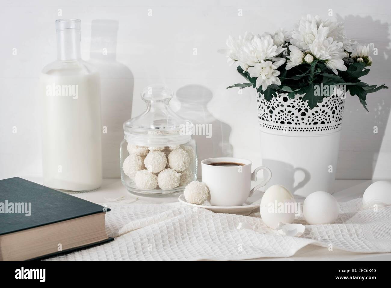 Flasche Milch, Buch, Kokos-Bonbons im Glas, Tasse Espresso-Kaffee, Blumentopf mit chrysanthen, Eier, Küchentuch auf dem Tisch. Stockfoto