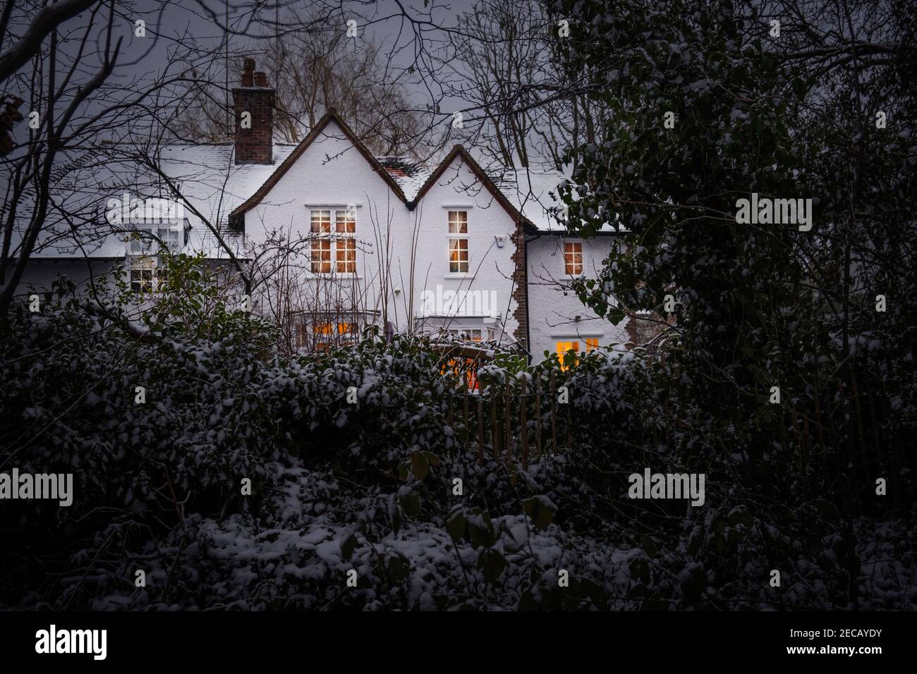 Ein Kunsthandwerk-Haus mit beleuchteten Fenstern, im Winter durch Waldbäume geschossen, Schnee auf dem Boden, Hampstead Garden Vorort, Barnett, Großbritannien Stockfoto