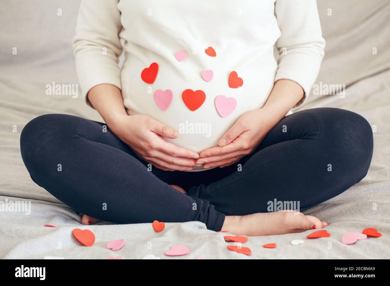 Nahaufnahme einer schwangeren Frau in weißen Kleidern, die auf dem Bett sitzt und ihren Bauch berührt. Schwangere Bauch mit rot rosa Papier Herzen bedeckt. Junge Dame erwartet Stockfoto