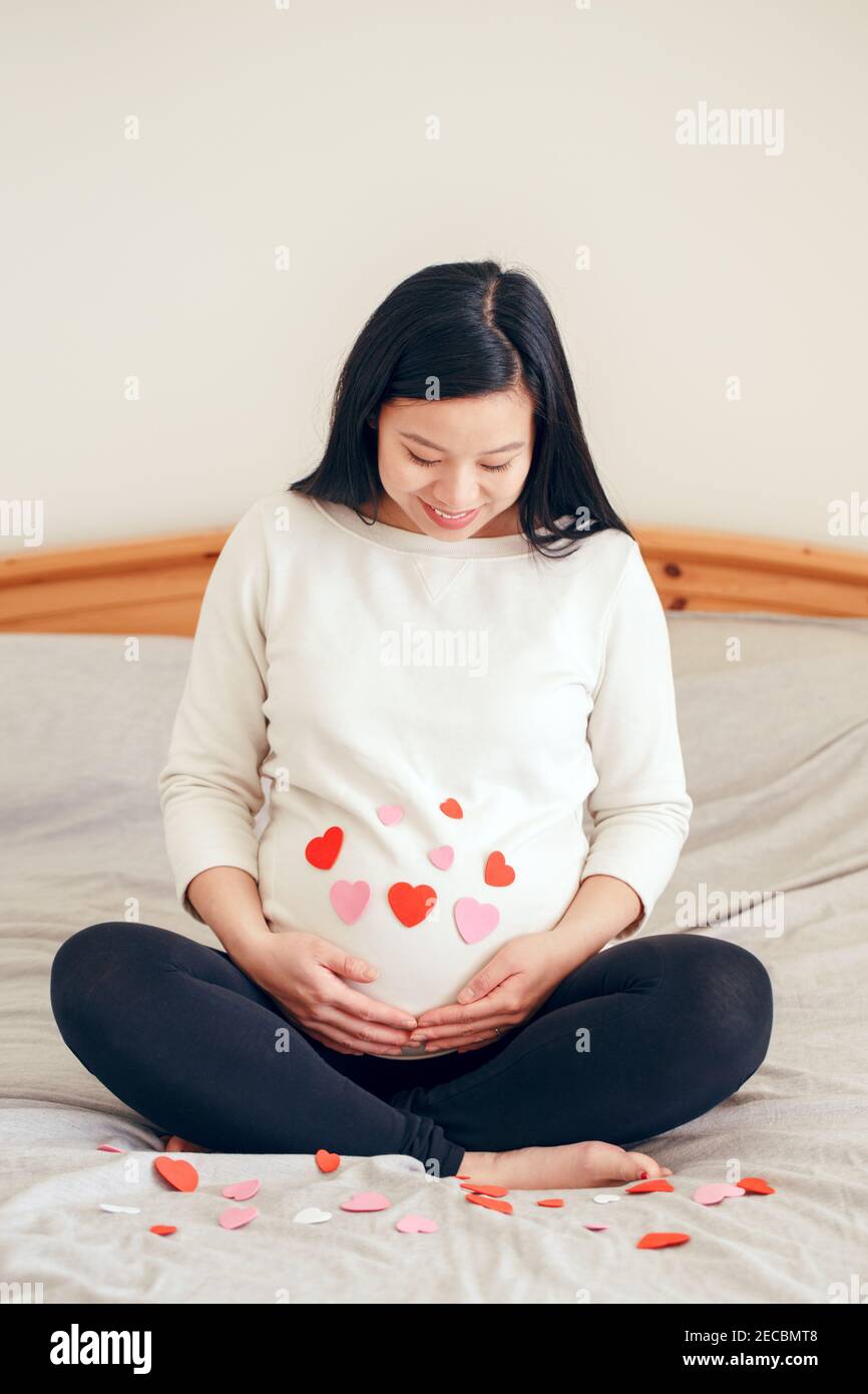 Lächelnd asiatisch chinesisch schwanger Frau sitzt auf Bett berühren ihren Bauch. Schwangere Bauch mit rot rosa Papier Herzen bedeckt. Junge Dame erwartet bei hom Stockfoto