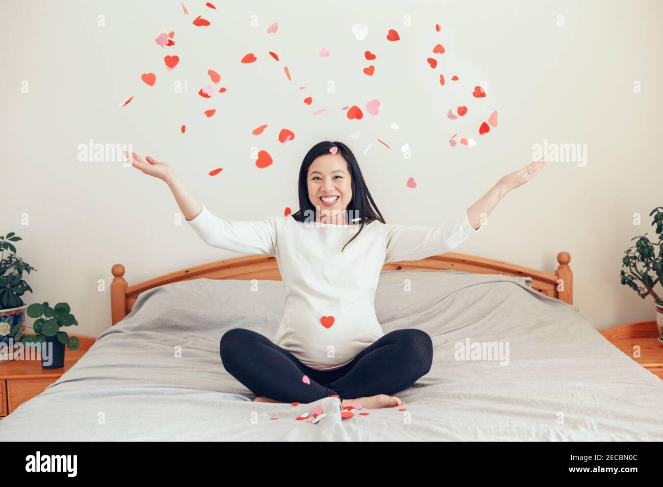 Lächelnde asiatische chinesische schwangere Frau auf dem Bett sitzen werfen kleine rote rosa Papier Herzen. Erwartet aufgeregt junge Dame weiblich zu Hause. Gesund glücklich p Stockfoto