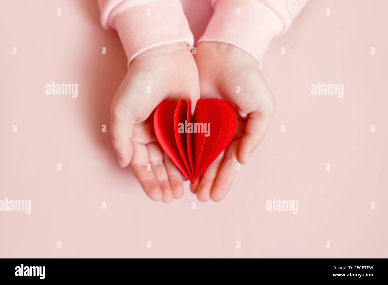 Nahaufnahme von Kind Kind Hände halten roten Papier gefaltet Herz auf hellrosa Hintergrund. Valentinstag Urlaub. Unterstützung, Pflege und Liebe Konzept. Rotes Herz Stockfoto