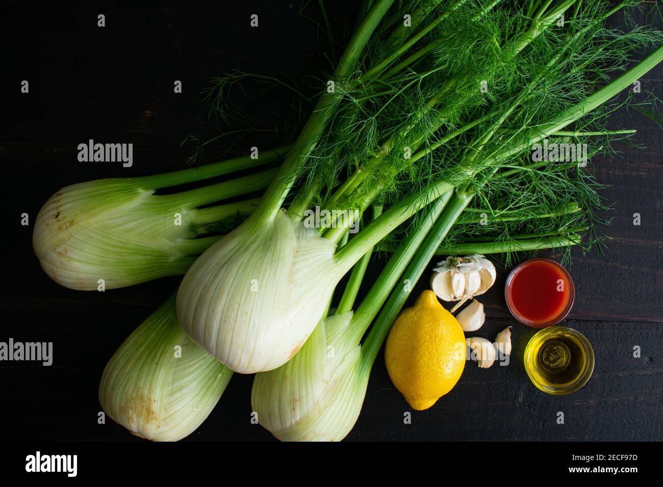 Sautierte Fenchel mit Knoblauch und Tomatensauce Zutaten: Rohe Fenchelzwiebeln, Zitrone, Knoblauch, natives Olivenöl extra und Tomatensaft auf dunklem Holz Stockfoto