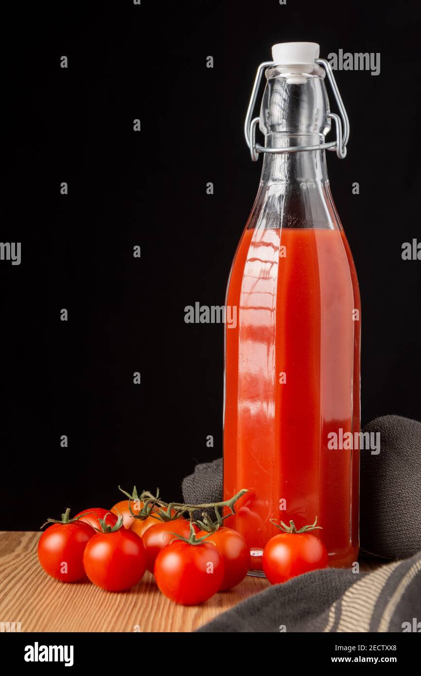 Ansicht der alten Glasflasche mit Tomatensaft, auf Holz mit Kirschtomaten, dunkles Tuch, schwarzer Hintergrund, vertikal Stockfoto