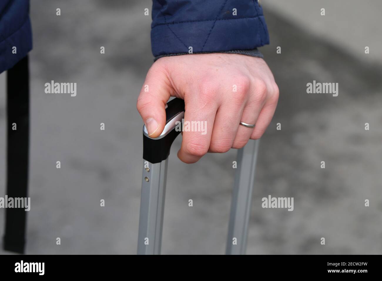 Kaukasischer Mann, der einen Koffergriff hält. Er hat einen Ehering und eine blaue, edle Jacke. Der Griff ist silber und schwarz. Nahaufnahme des Farbbildes. Stockfoto